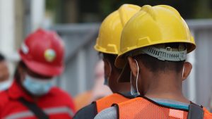 Fotografiet viser to arbeidere med hjelm, sikkerhetsvest og munnbind som venter på å bli kontrollert av en tredje mann.