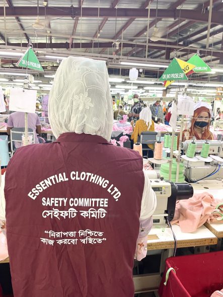 Person står med ryggen til i tekstilfabrikk, har på seg en vest med teksten Safety Committee 
